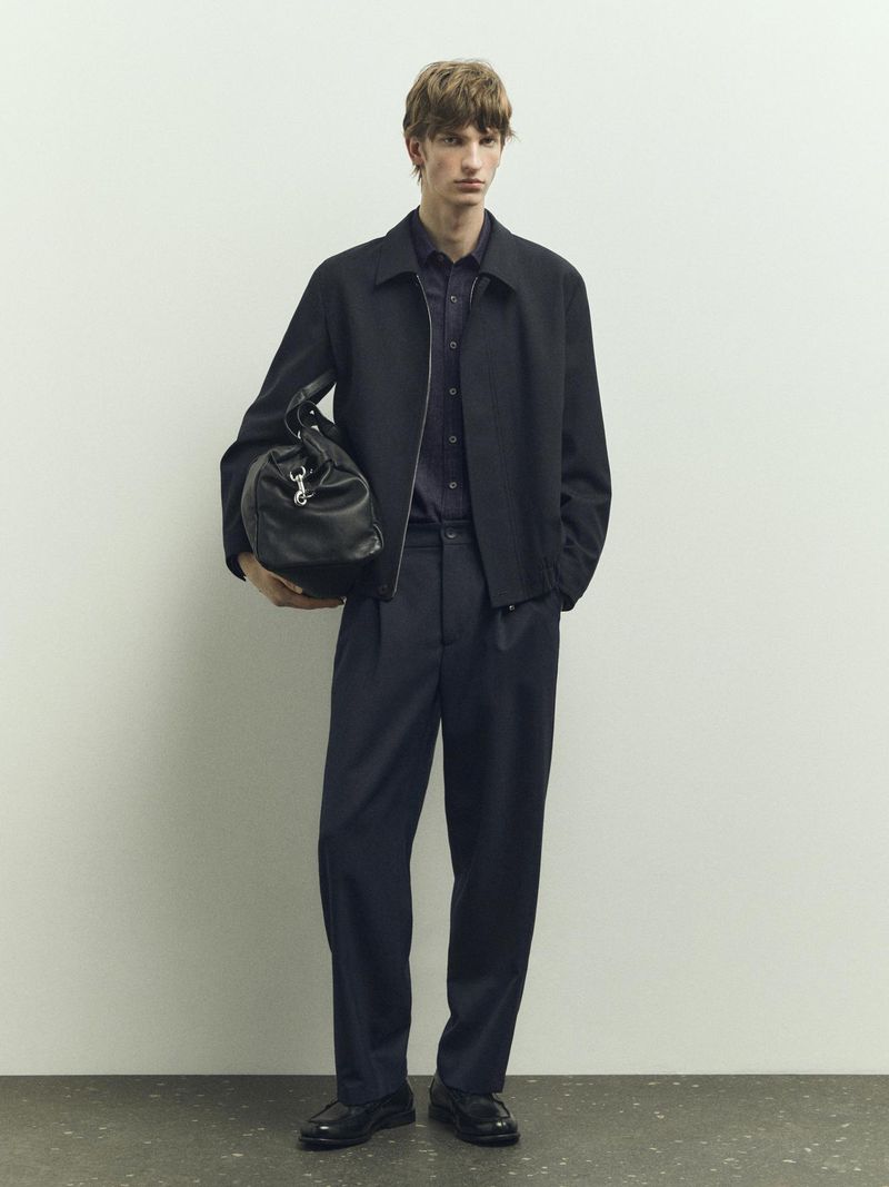 A fair-skinned male model in a navy shirt, jacket, and pants holds a black leather duffel bag in a minimalist studio setting.
