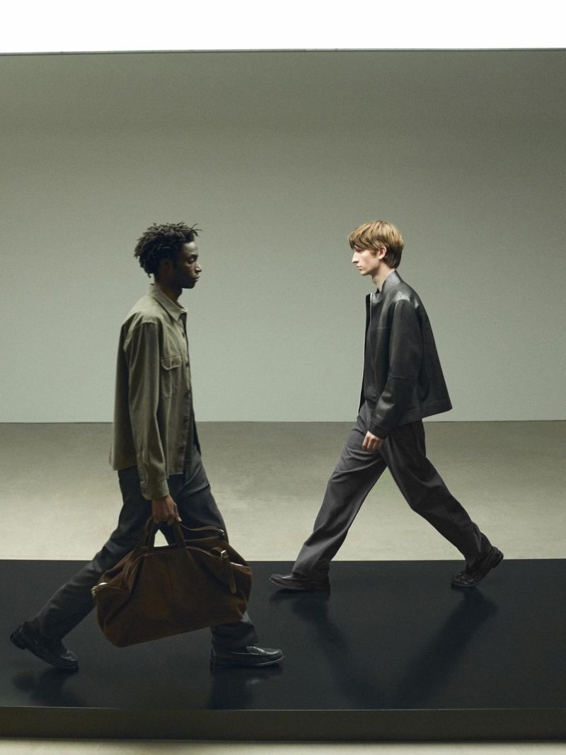 Two men in stylish outfits walk past each other in a studio setting. One man carries a large leather duffel bag, horizontal studio shot.