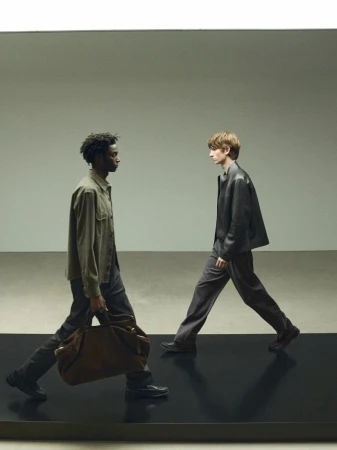 Two men in stylish outfits walk past each other in a studio setting. One man carries a large leather duffel bag, horizontal studio shot.