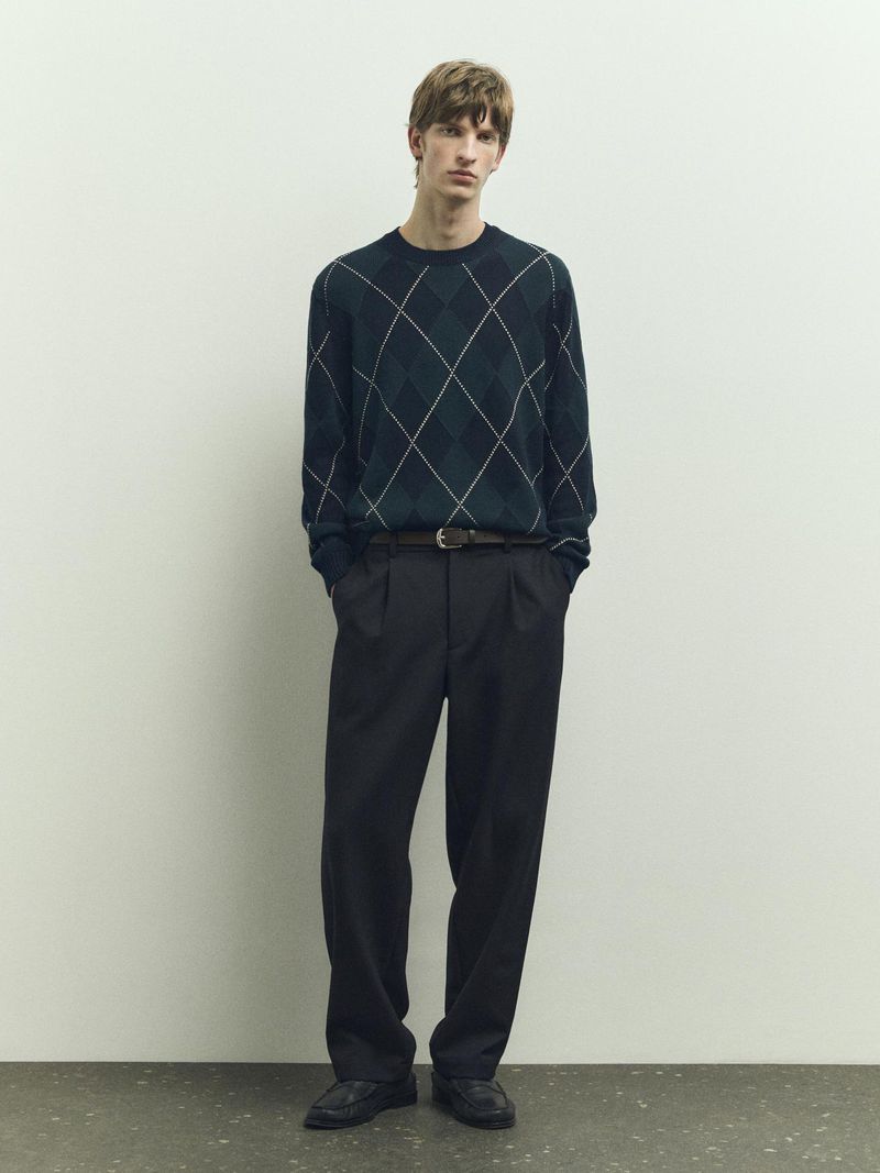 Full-length studio shot of an adult male wearing an argyle patterned sweater, black trousers, a belt, and black shoes. He stands in front of a white wall.