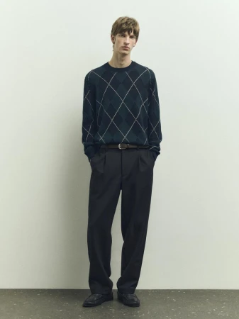 Full-length studio shot of an adult male wearing an argyle patterned sweater, black trousers, a belt, and black shoes. He stands in front of a white wall.