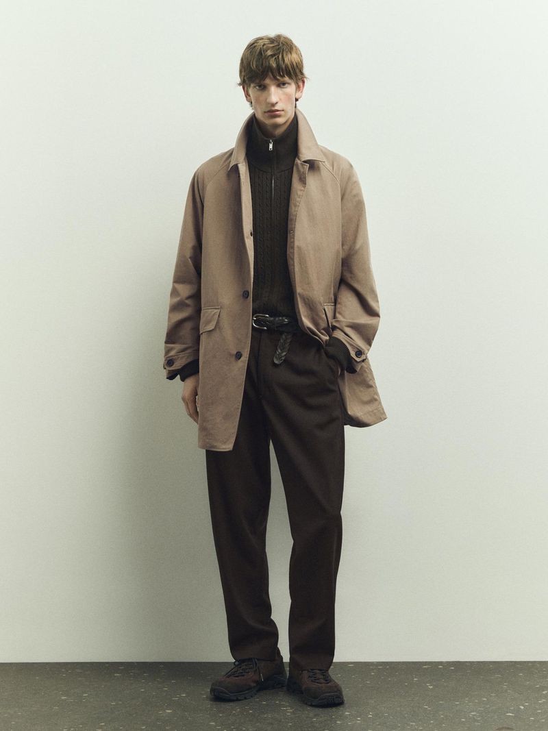 A man in a tan overcoat stands in front of a pale wall. He is wearing dark trousers, a zip-up sweater, and brown shoes in a studio shot.