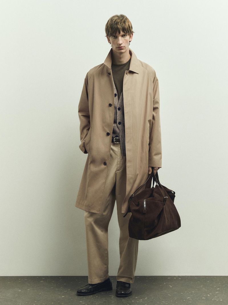A male fashion model poses indoors wearing a trench coat, pants, sweater vest, and leather bag in a studio setting on a gray background.
