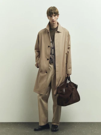 A male fashion model poses indoors wearing a trench coat, pants, sweater vest, and leather bag in a studio setting on a gray background.