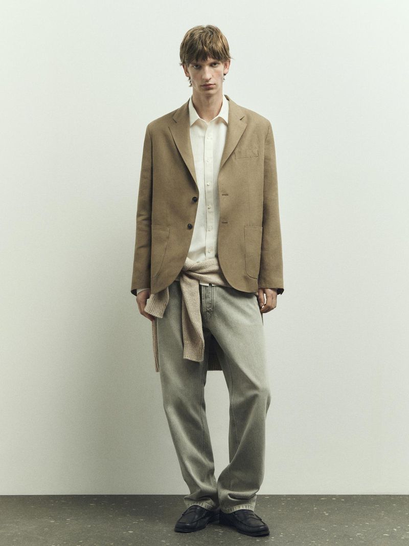Full-length shot of a slender adult man wearing a blazer, button-down shirt, jeans, and loafers, posing in front of a light-colored wall.