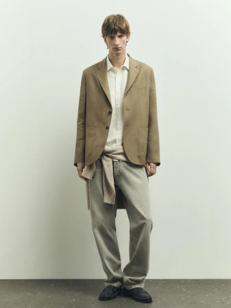 Full-length shot of a slender adult man wearing a blazer, button-down shirt, jeans, and loafers, posing in front of a light-colored wall.