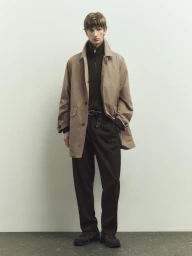 Full-length portrait of a male model wearing a camel coat over a dark zip-up sweater and trousers against a pale wall.