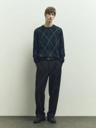 Model wearing dark argyle sweater and trousers standing against a light seamless studio backdrop for Massimo Dutti PS 026 Collection