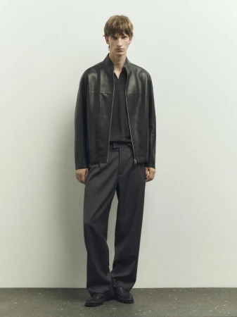 A male model wearing a dark leather jacket and grey trousers standing against a neutral studio background.