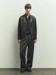 A male model wearing a dark leather jacket and grey trousers standing against a neutral studio background.