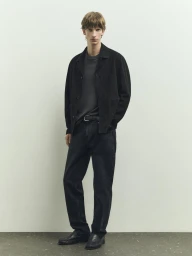 Full length shot of a young man wearing a dark jacket, grey t-shirt, and dark trousers standing against a pale seamless studio background.