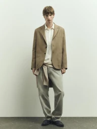 Full-length portrait of a male model wearing a brown blazer, white shirt, and grey trousers against a seamless neutral background.