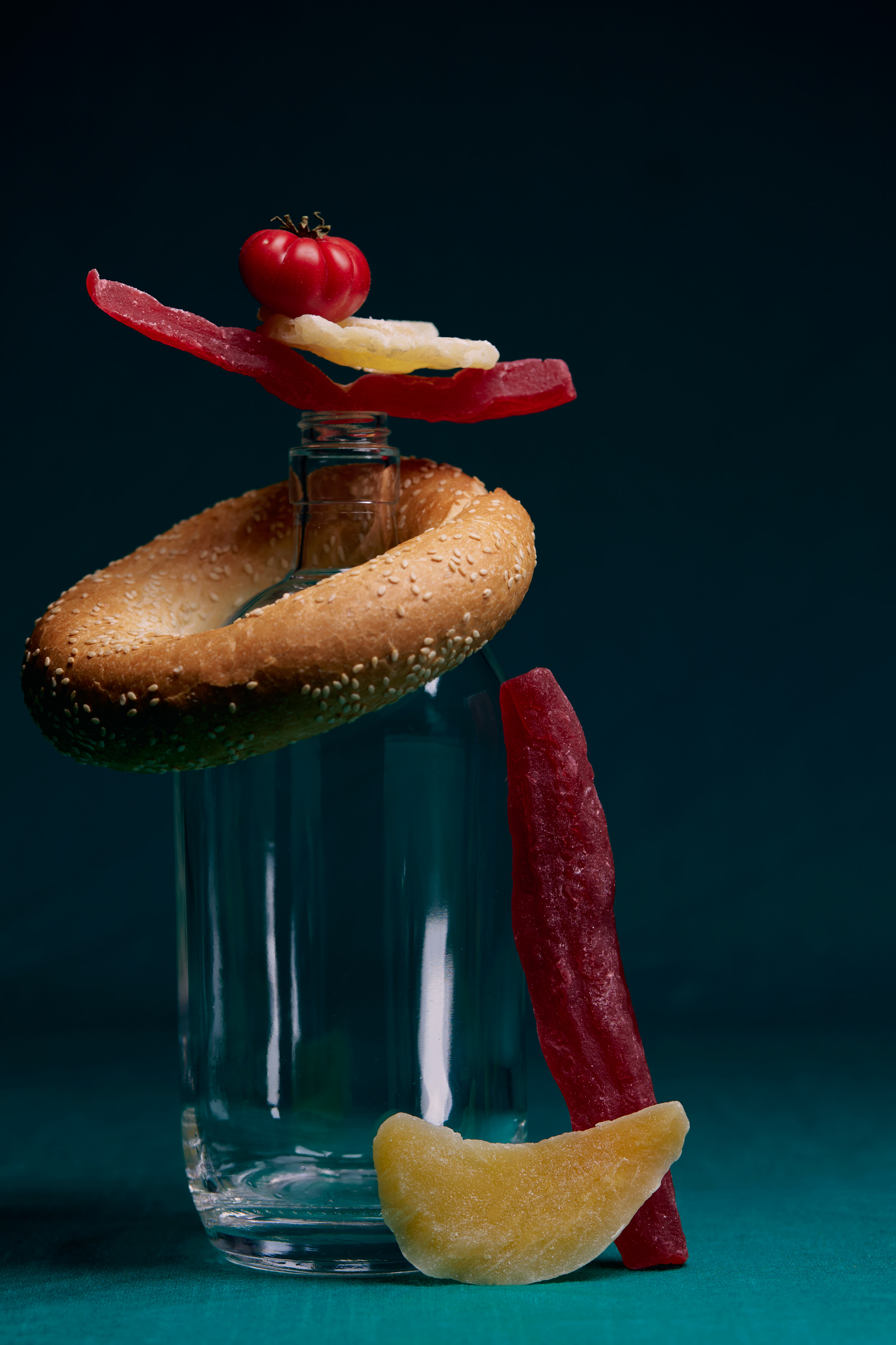 Still life food photography featuring a bagel, tomato, and dried fruits balanced on a clear glass bottle against a dark background.