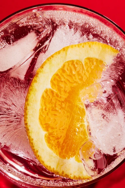 Close-up top shot of a cocktail glass filled with ice cubes, an orange slice, and a sparkling beverage against a vibrant red background.