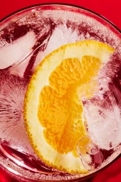 Top-down view of a glass filled with carbonated beverage, ice cubes, and an orange slice against a solid red background
