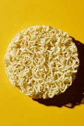 Overhead studio shot of a dry round instant noodle block centered on a solid yellow background with casting shadows.