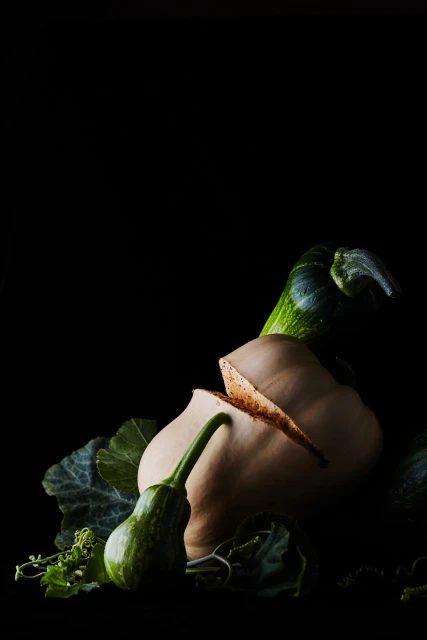 Still life of butternut squash and green gourds with leaves arranged on a black backdrop, creating a dramatic contrast.