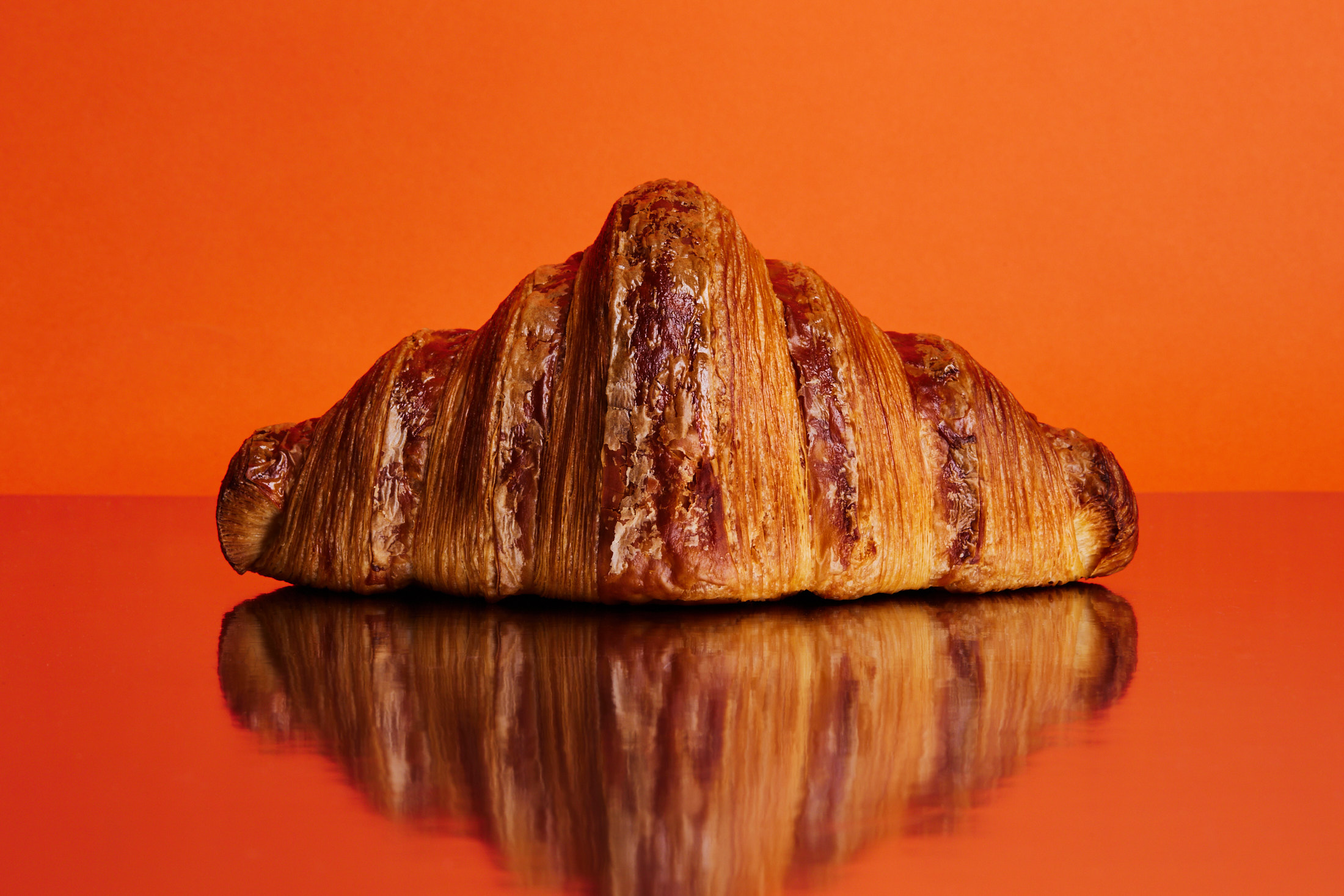Close-up studio shot of a glossy croissant on a reflective orange surface, set against a vibrant orange background.