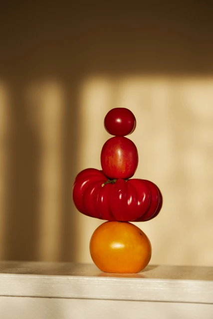 Stack of red and yellow heirloom tomatoes balanced on a white surface against a cream-colored wall with window shadow.