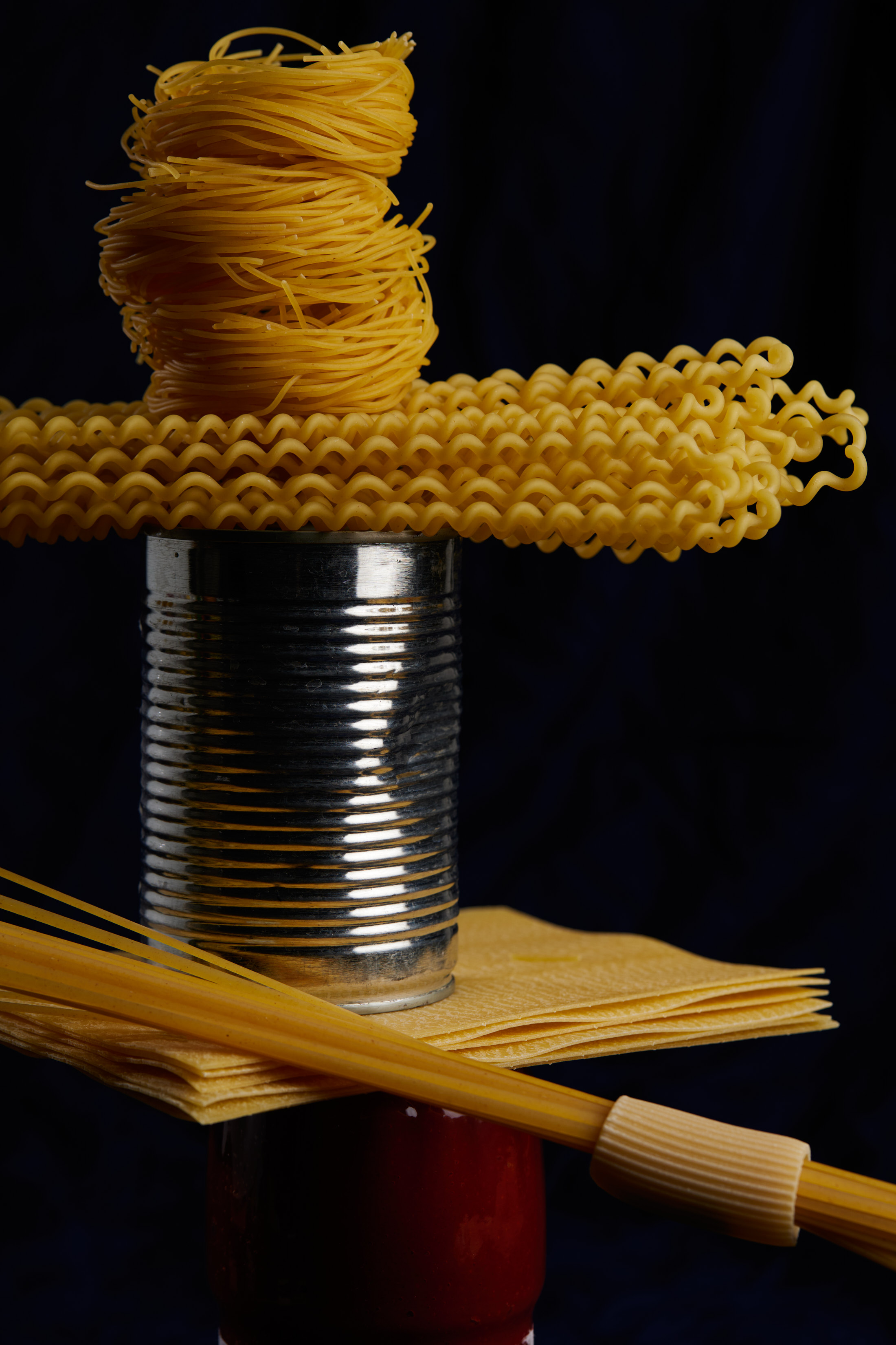 Arrangement of fusilli lunghi, spaghetti, tomato sauce jar, and a silver can on a dark background, food photography, product photography.