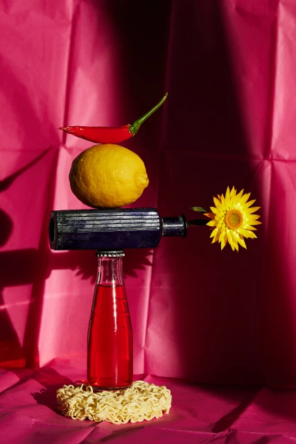 Still life photo of a stacked arrangement with a red pepper, lemon, metal pipe, drink bottle, and flower against a pink backdrop.