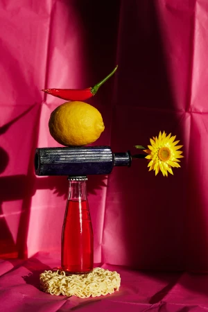 Still life photo of a stacked arrangement with a red pepper, lemon, metal pipe, drink bottle, and flower against a pink backdrop.