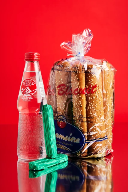 A glass bottle of Uludag mineral water, two green chocolate bars, and a bag of Eti Biscuit are against a shiny red background.