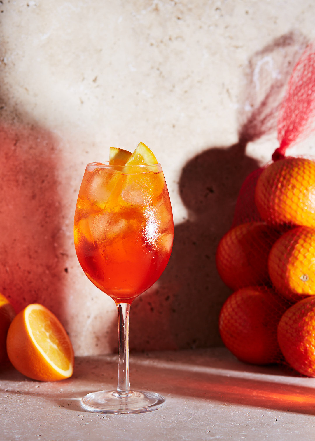 Aperol spritz in wine glass with orange slices and ice cubes; oranges in mesh bag on table with reddish and grey background.