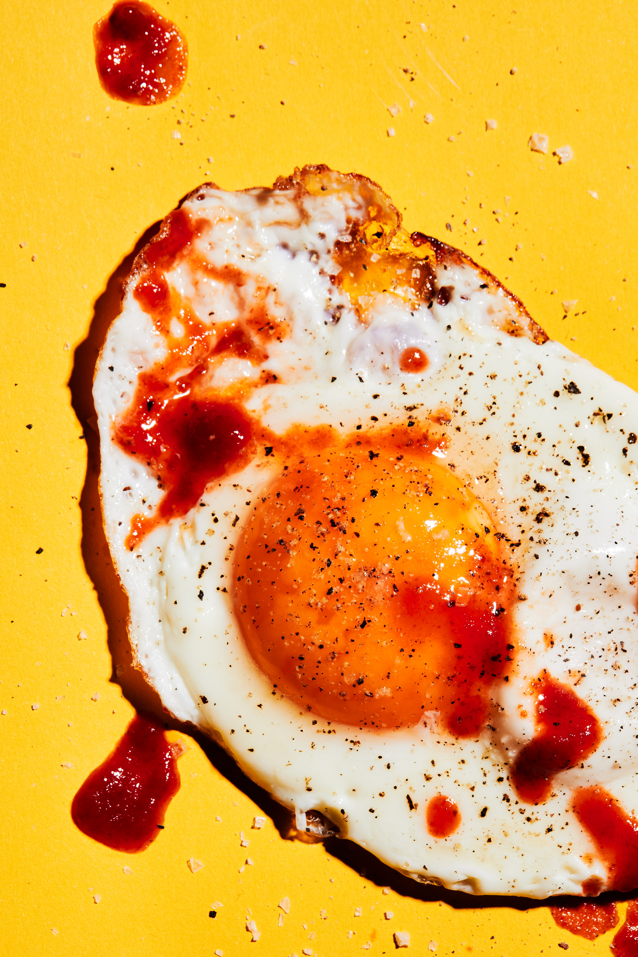 Overhead view of a fried egg with bright yellow yolk and pepper seasoning on a yellow background, some tomato sauce on the egg and background.