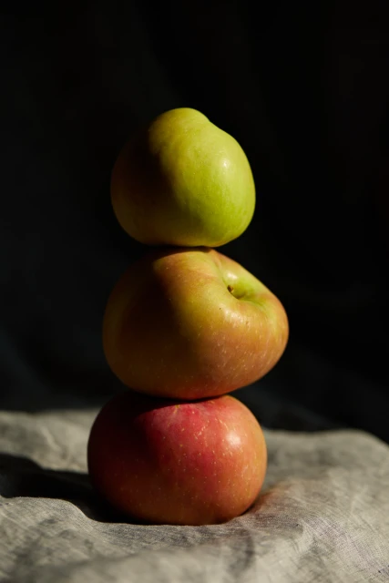 A still life photograph of three apples stacked on top of each other on a wrinkled gray cloth against a dark background, lit from above.