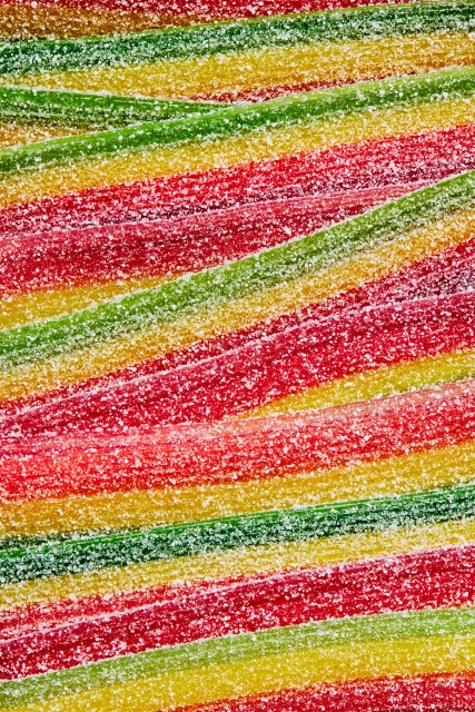 Overhead close-up of colorful, sugar-coated sour candy belts in horizontal rows. Red, green, and yellow stripes create a vibrant, sugary texture.