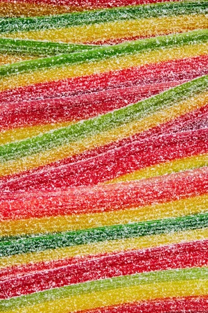 Overhead close-up of colorful, sugar-coated sour candy belts in horizontal rows. Red, green, and yellow stripes create a vibrant, sugary texture.