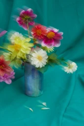 A vase of colourful flowers captured with motion blur against a textured teal fabric background by Rebecca Newman.