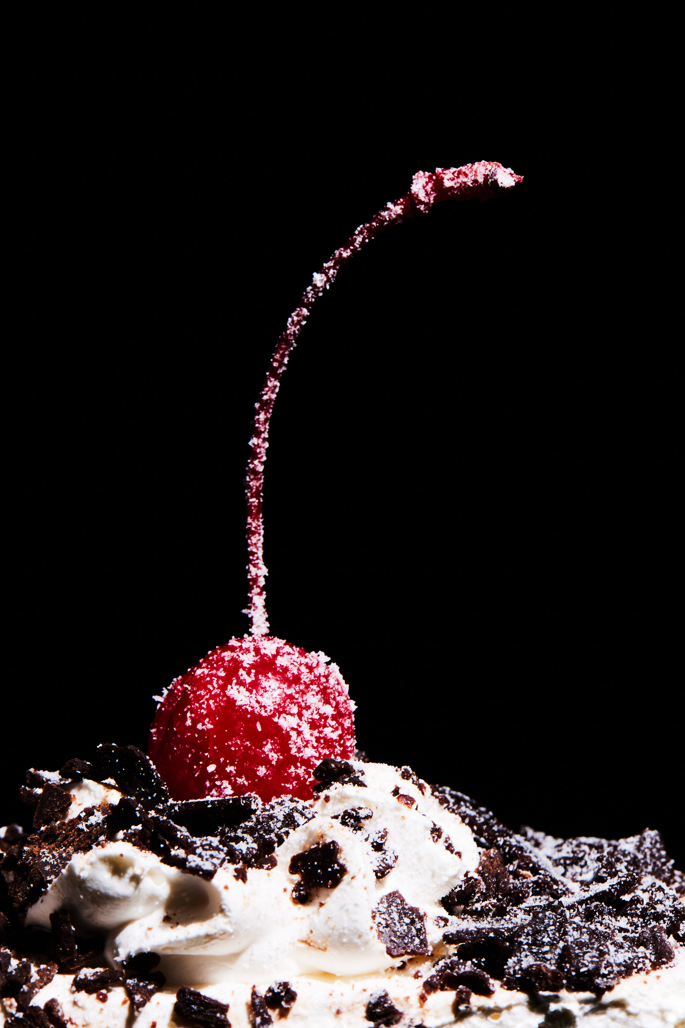 Close-up of a red maraschino cherry dusted with sugar, atop white whipped cream and dark chocolate shavings, against a black backdrop.