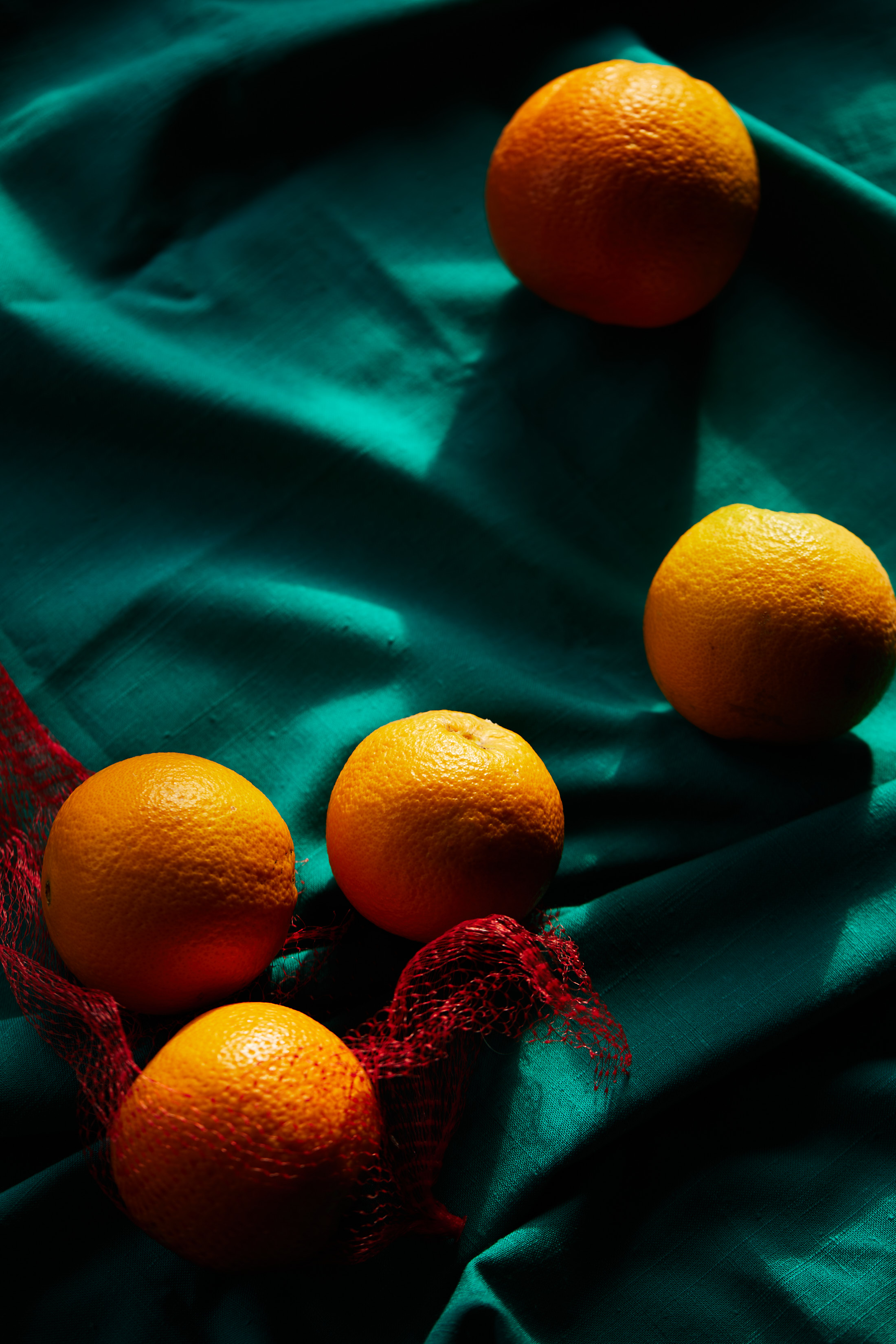 Still life featuring a cluster of oranges in a net bag with other oranges scattered on a wrinkled turquoise textile background, in shadow.