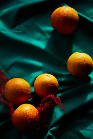 Still life featuring a cluster of oranges in a net bag with other oranges scattered on a wrinkled turquoise textile background, in shadow.