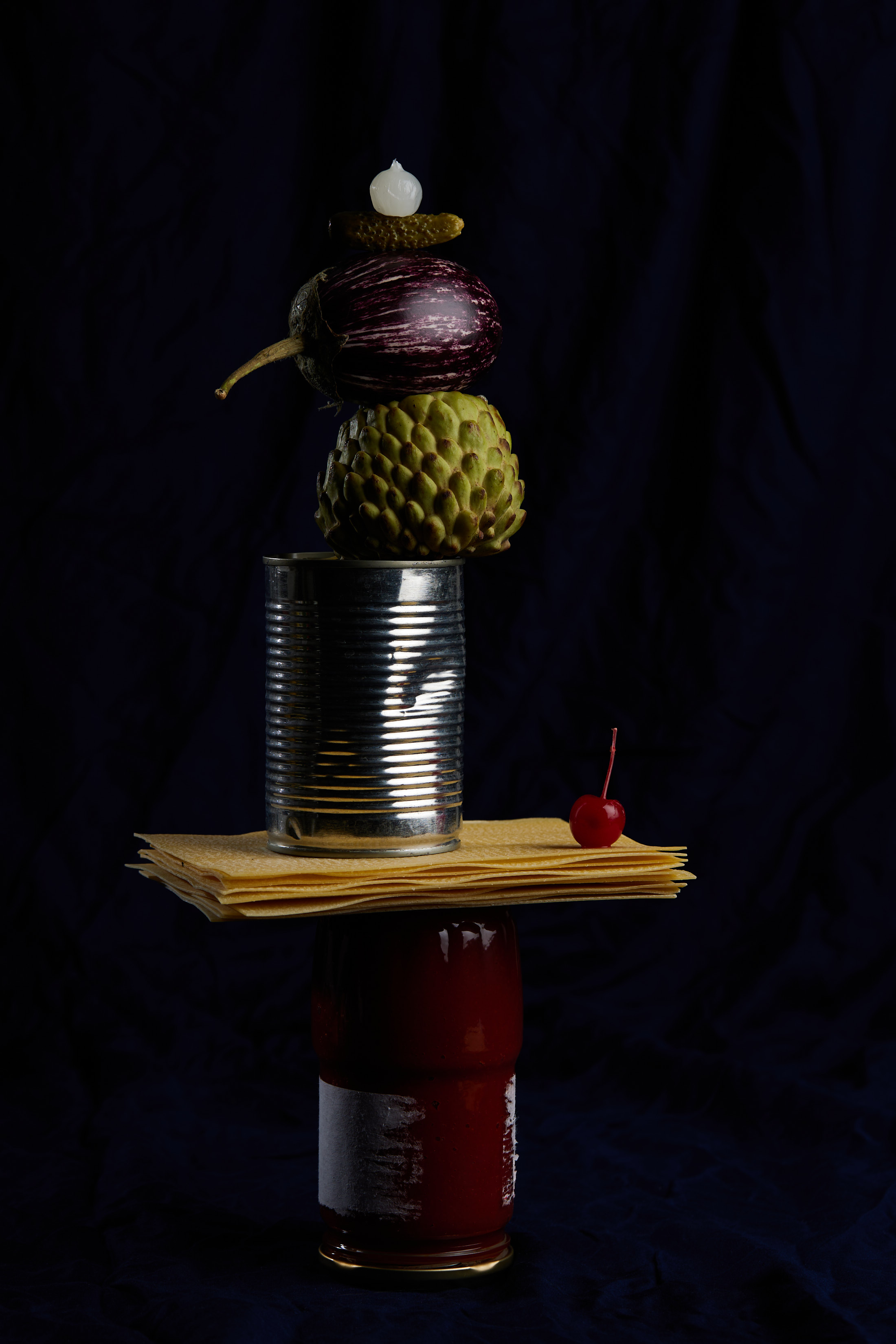 Still life food photography of stacked food items: cans, lasagna noodles, eggplant, cherimoya, pickle, against a dark background.