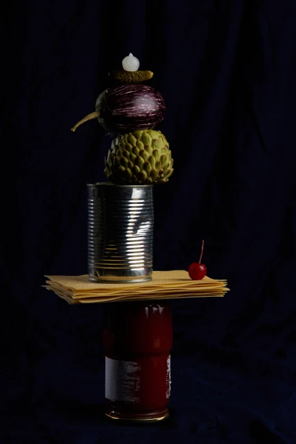 Still life food photography of stacked food items: cans, lasagna noodles, eggplant, cherimoya, pickle, against a dark background.