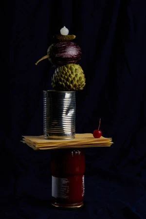 Still life food photography of stacked food items: cans, lasagna noodles, eggplant, cherimoya, pickle, against a dark background.