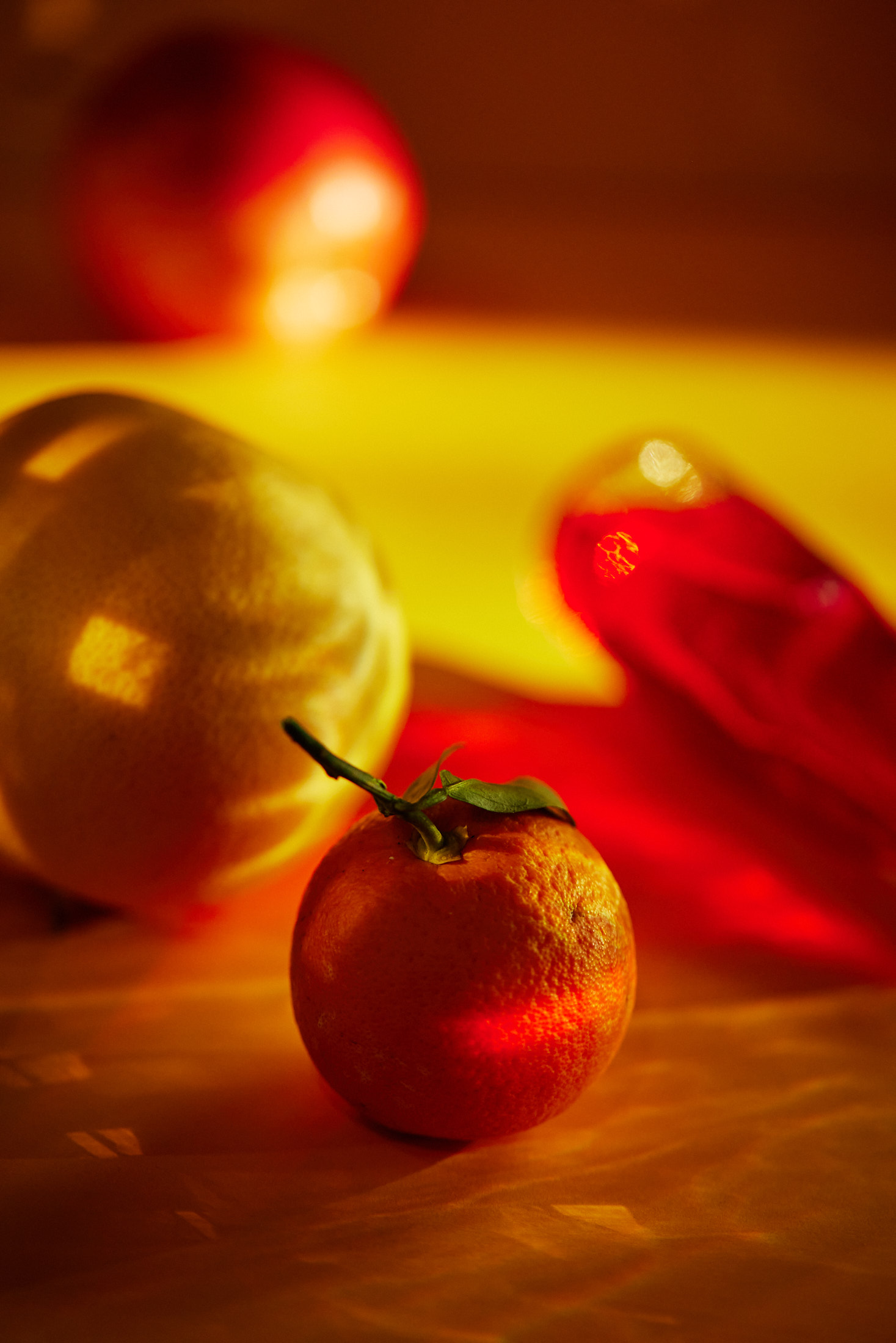 Still life photograph featuring an orange in the foreground, with a lemon, peach, and carafe of red liquid blurred in the background.