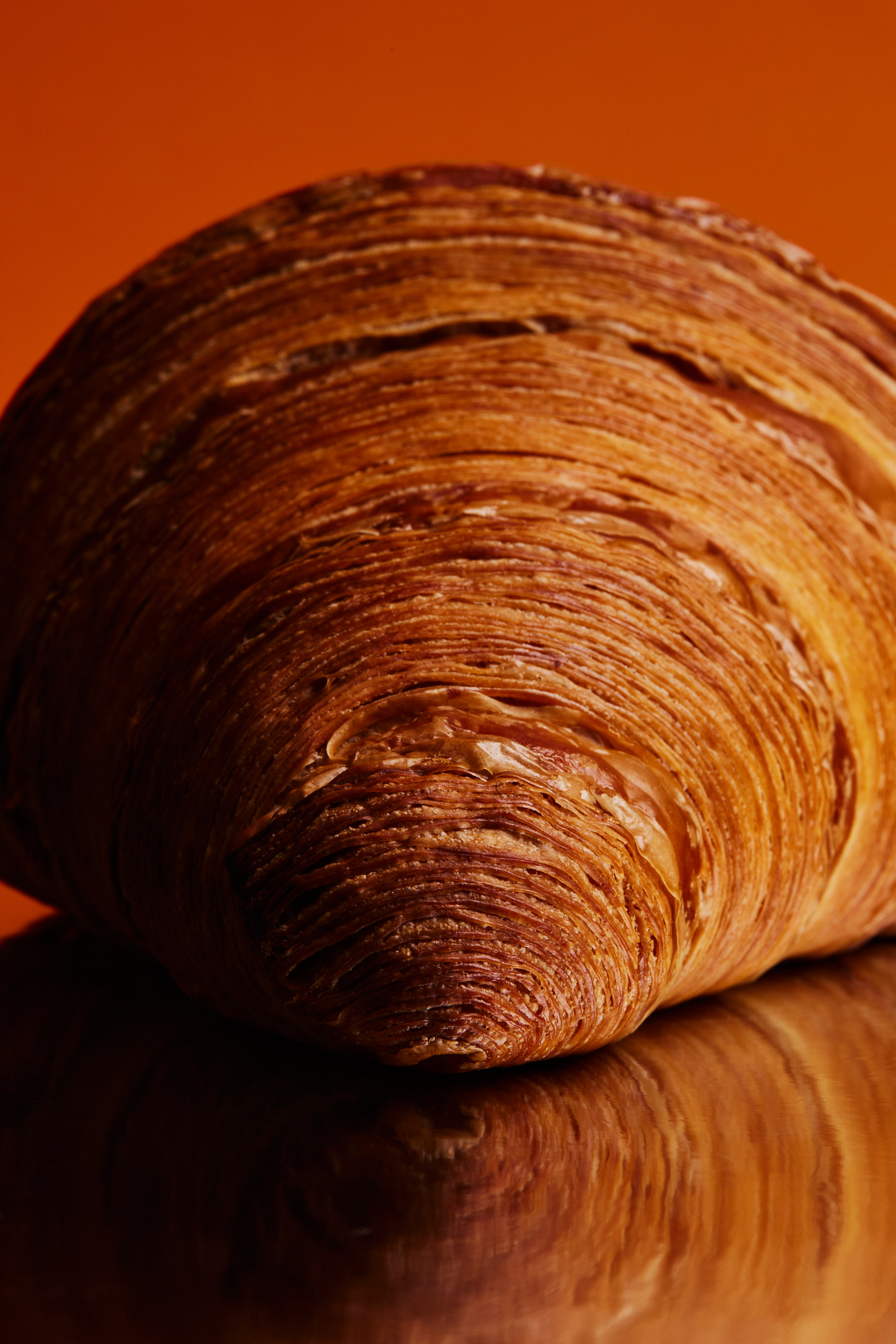 Close-up studio shot of a golden-brown croissant with layered texture, resting on a reflective surface against an orange background.