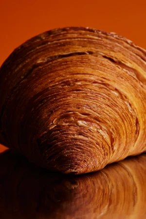 Close-up studio shot of a golden-brown croissant with layered texture, resting on a reflective surface against an orange background.