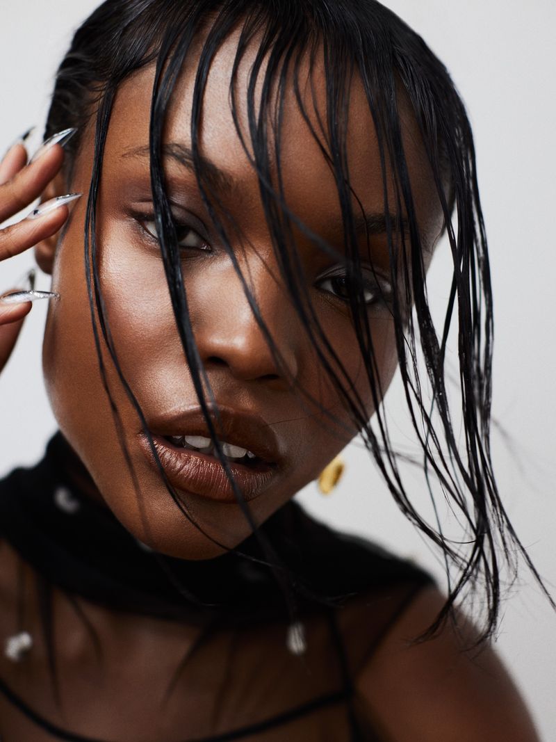 Close-up beauty shot of a Black woman with wet black hair styled across her forehead, glossy lips, wearing a sheer, beaded top on white.