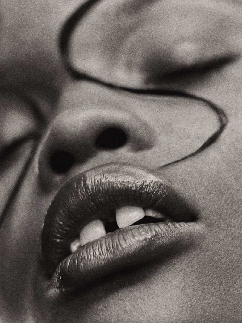 Monochrome close-up of a person's face featuring lips, teeth, nose, closed eyes, and a strand of hair against a neutral background.