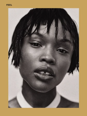 Monochrome portrait of a young Black woman with wet short black hair, gazing at the camera against a beige-grey background and white top.