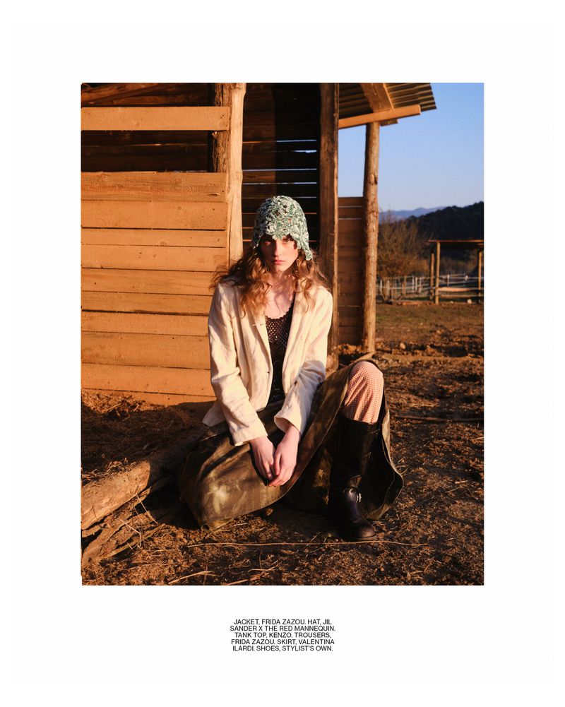 Woman sits in front of a rustic wooden shed, wearing a patterned bucket hat, jacket, and loose skirt, with a rural landscape background.