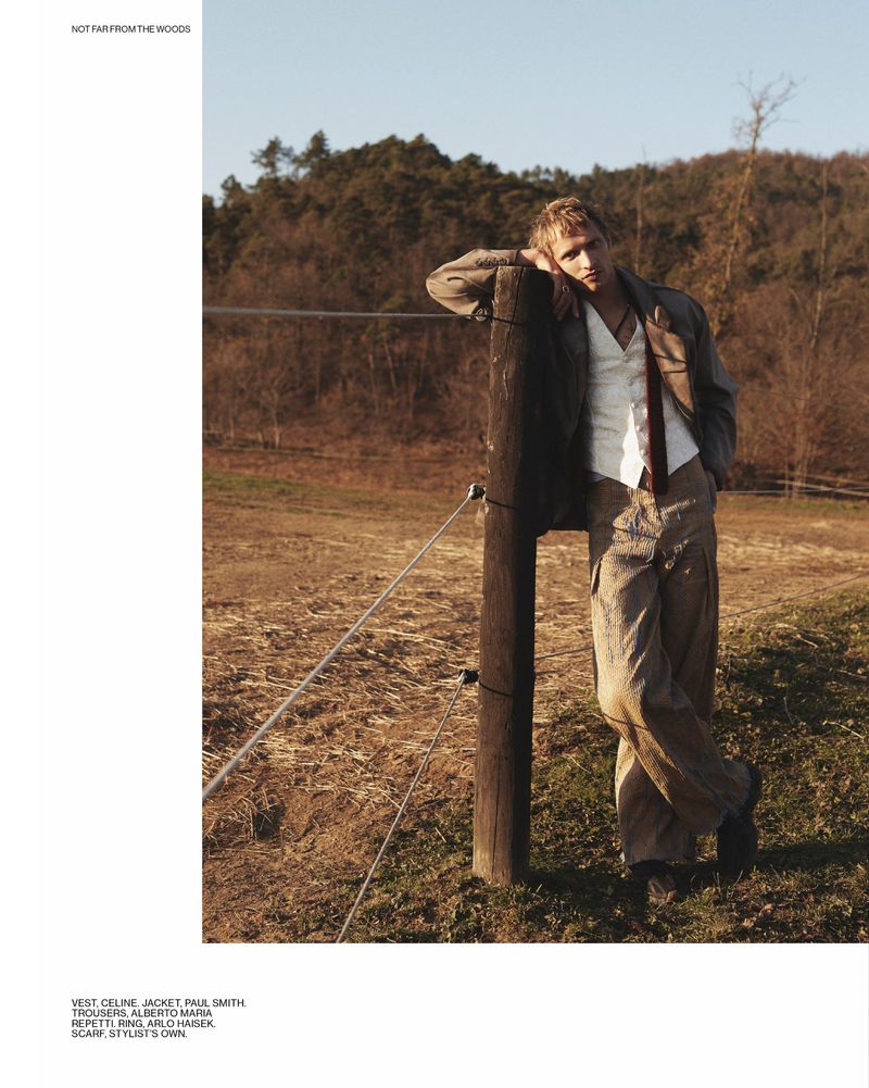 A blond male model in a jacket and vest leans against a wooden fence post in an outdoor fashion editorial with a grassy, wooded setting.