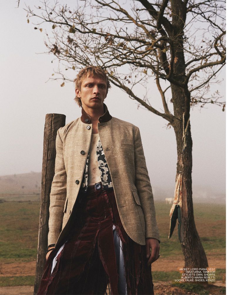 Man in stylish blazer and draped skirt stands outdoors near a tree, showcasing fashion in a natural setting with soft lighting.
