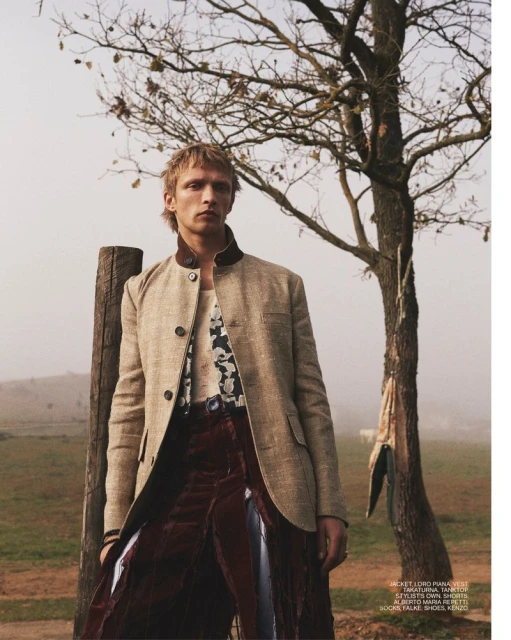 Man in stylish blazer and draped skirt stands outdoors near a tree, showcasing fashion in a natural setting with soft lighting.
