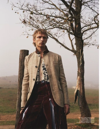 Man in stylish blazer and draped skirt stands outdoors near a tree, showcasing fashion in a natural setting with soft lighting.
